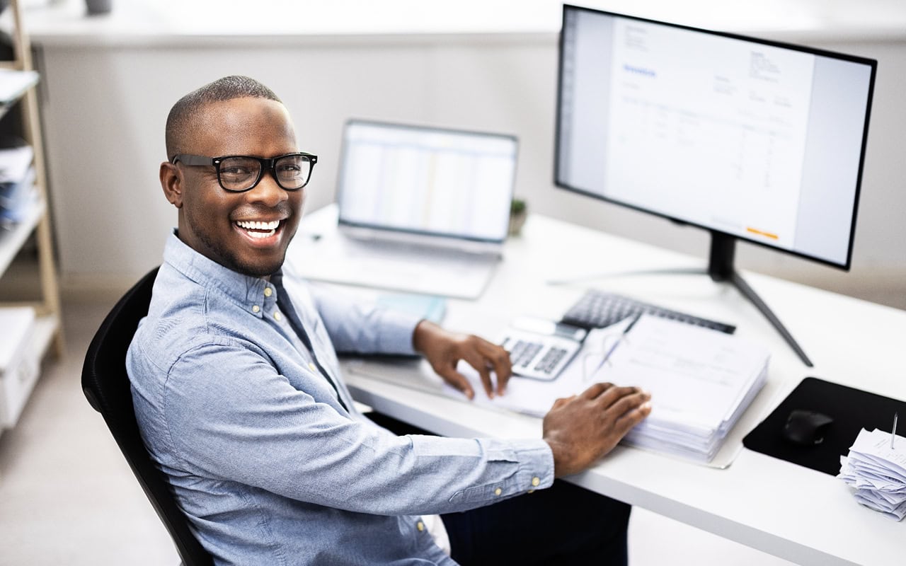 A smiling black man sitting at a desk with a computer.