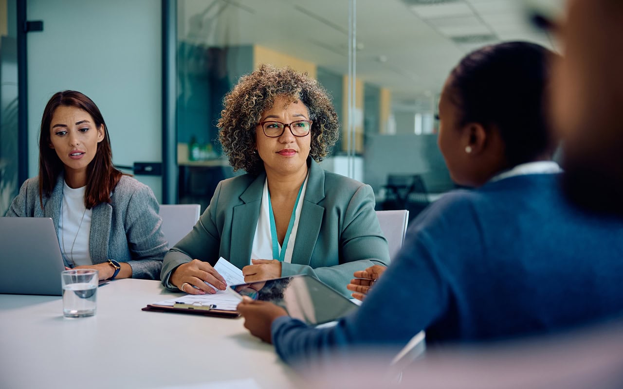Three businesswomen sitting at a table in an office. One is facing the other two with her back turned to the camera