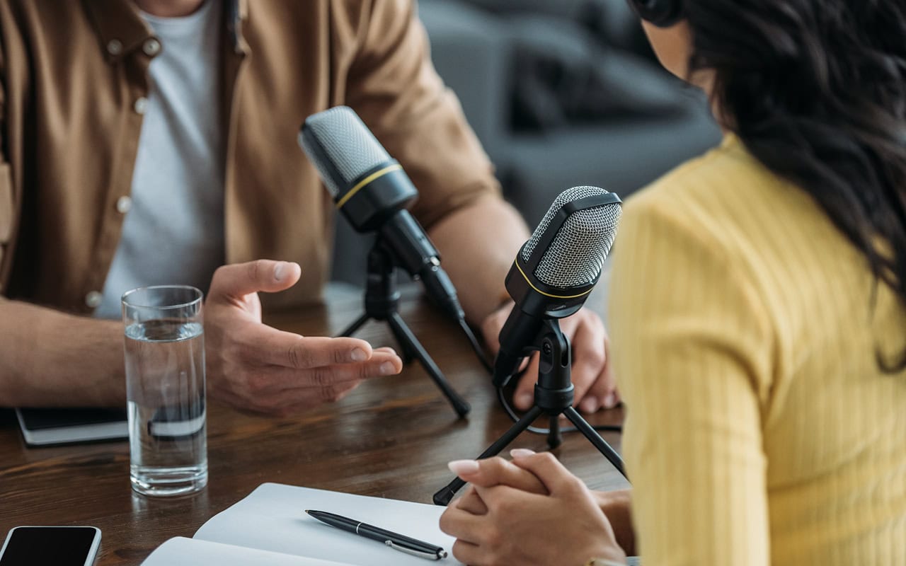 A man and woman sitting at a table with microphones conversing