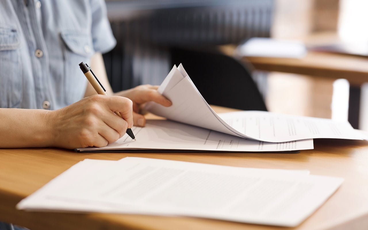 A person is writing on paper with a pen. The desk has more paper documents.