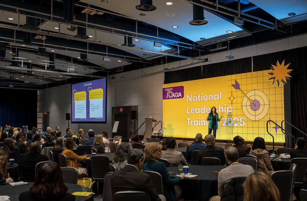 Image of an auditorium with a lot of people sitting facing a stage with a big yellow graphic branded for the event. On the stage is a female speaker.