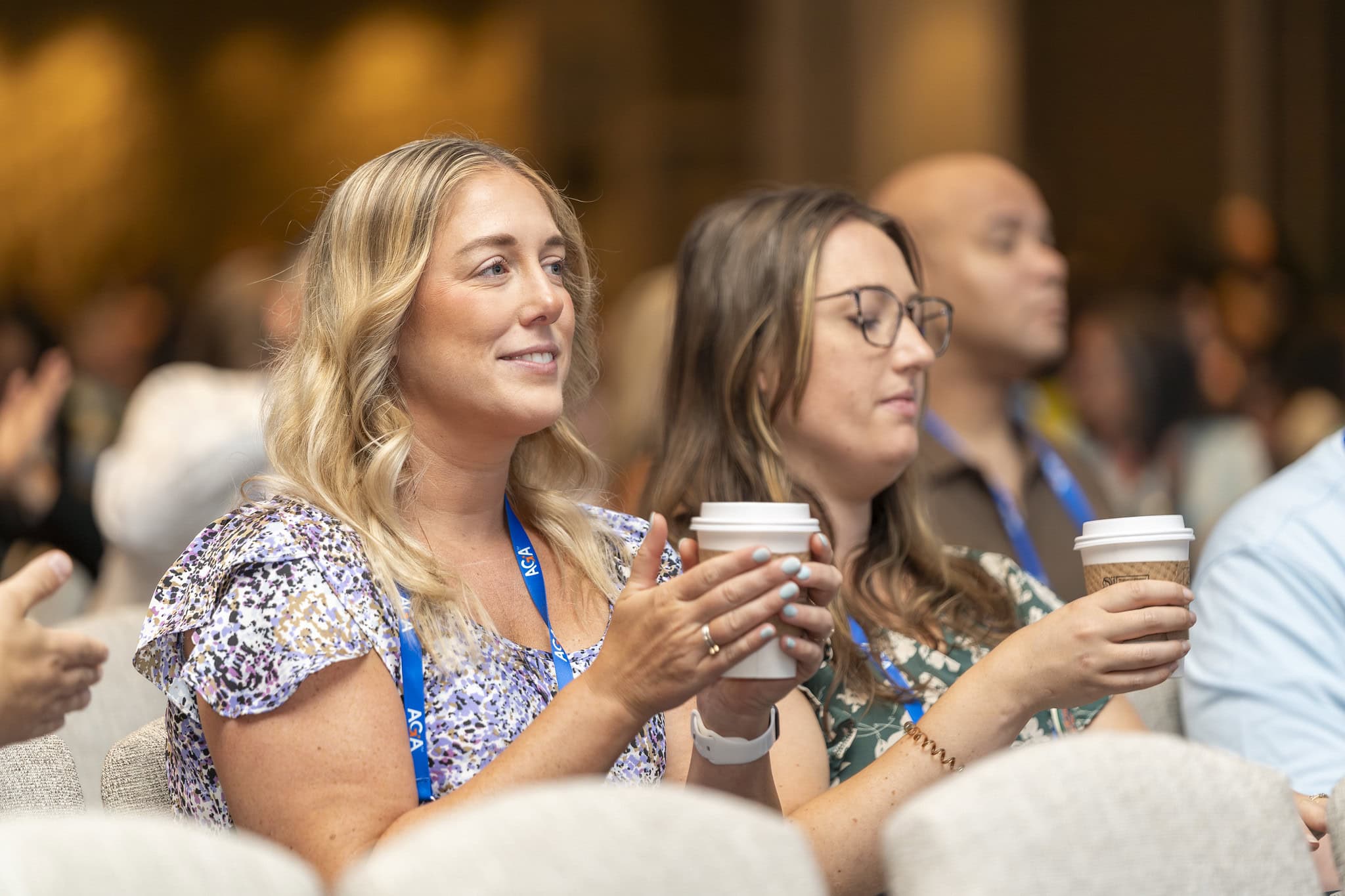 Two young women sit in the crowd at a training event holding coffee cups.