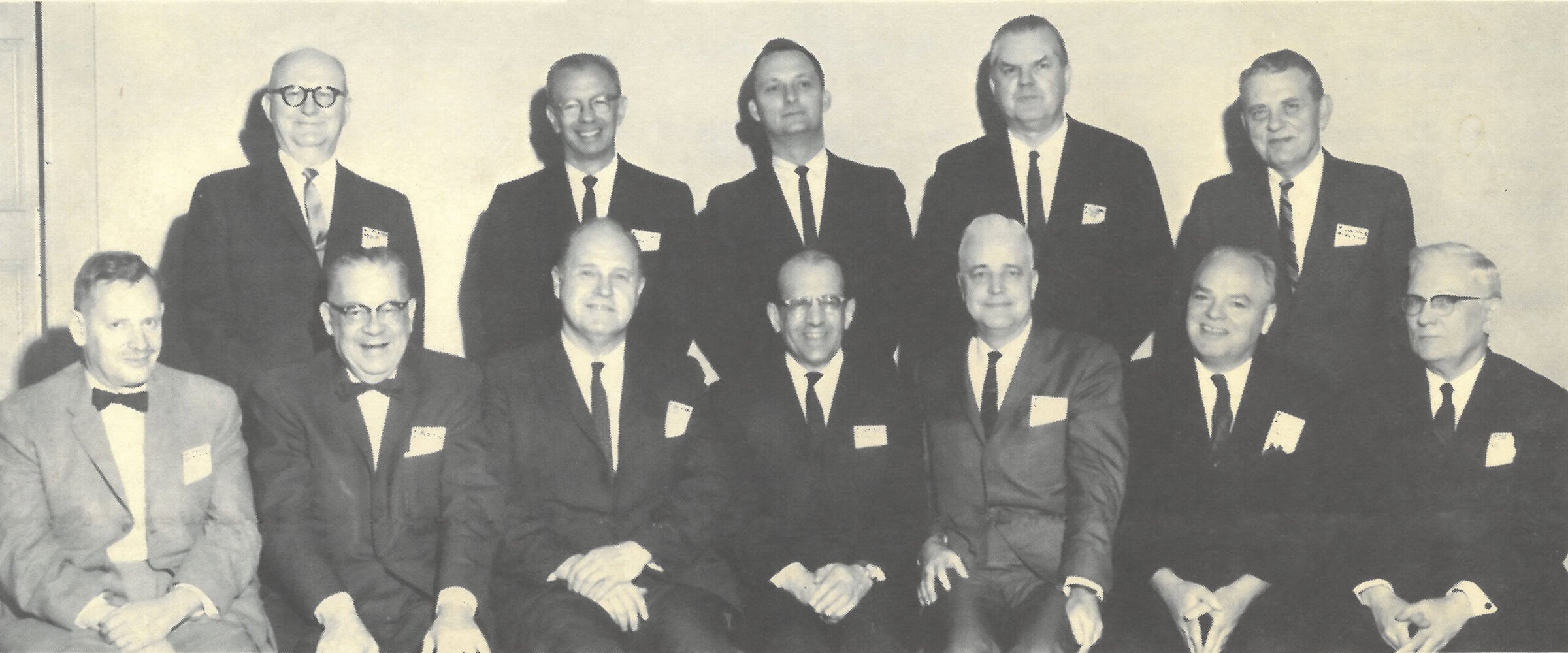 A black and white photo of a group of 12 dapper men in suits; seven men sit in the front row and five men stand in the second.