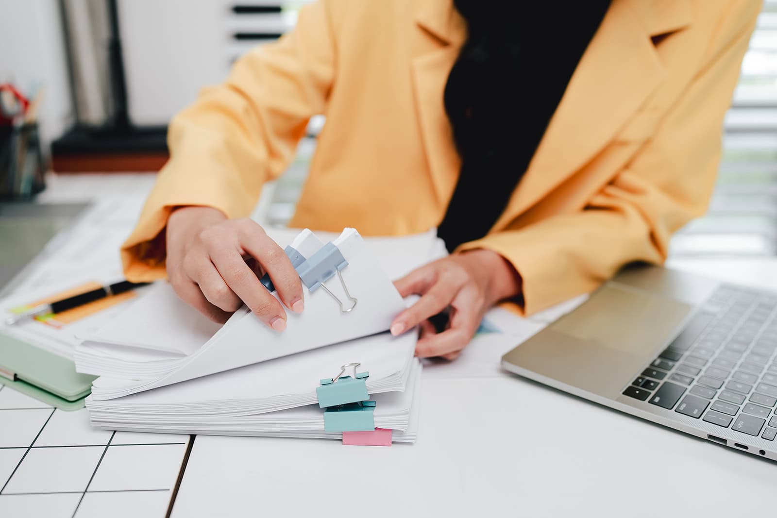 A person in a yellow jacket sits at a desk flipping through a stack of papers with sticky notes tabbing the edges